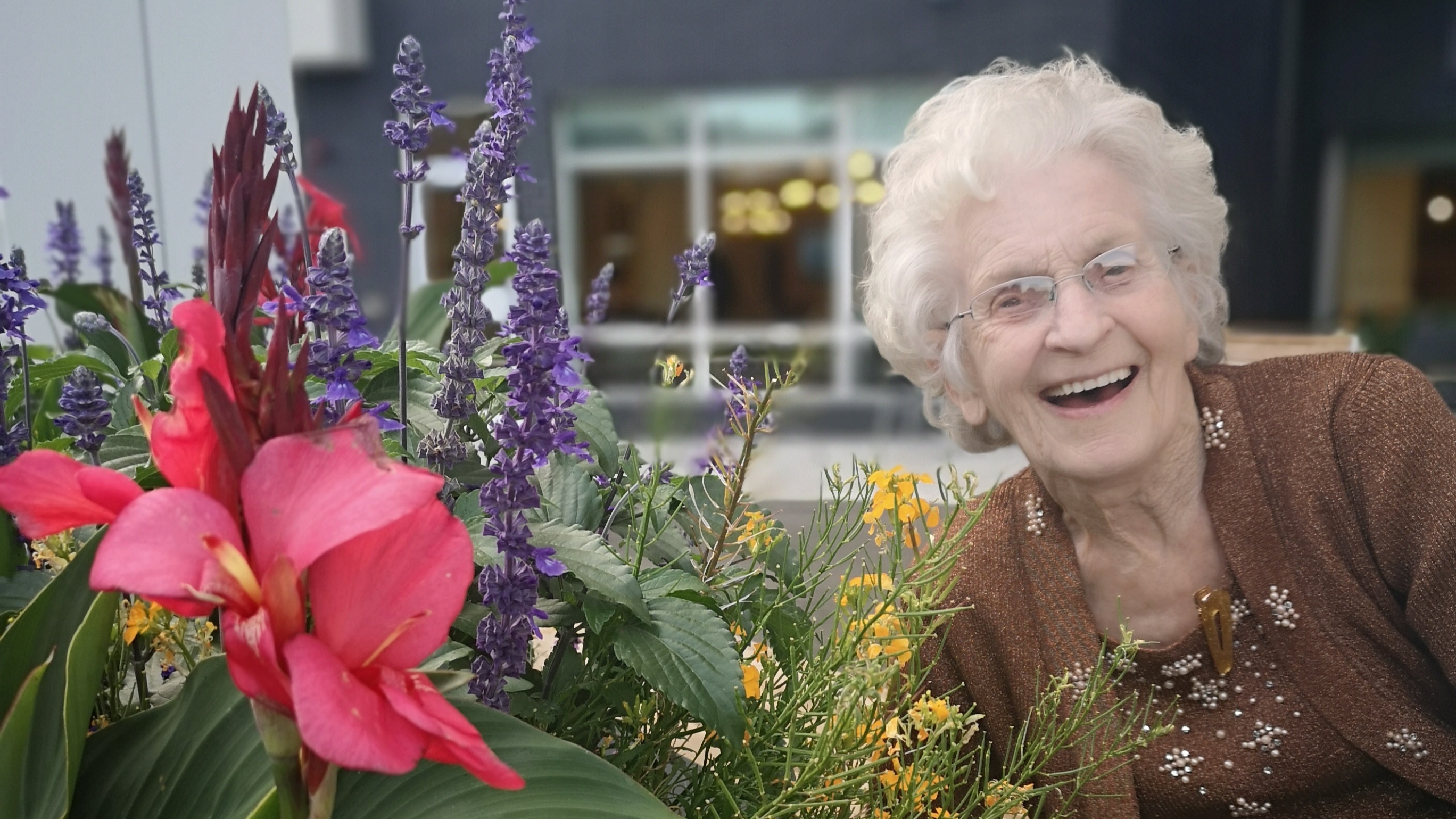 A senior lady smiling beside pink, purple and yellow flowers