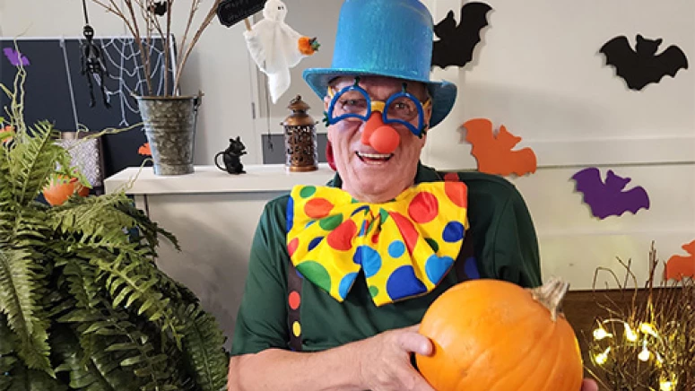Resident dressed in a clown costume for Halloween. He is smiling and holding a pumpkin.