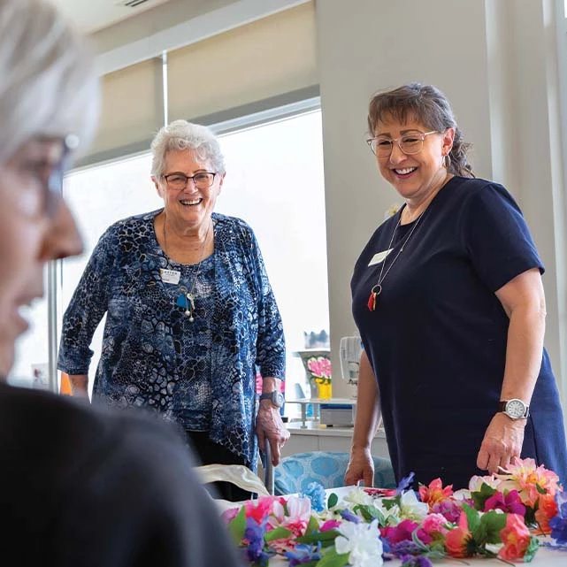 Senior woman laughing and smiling with an Optima Living staff member. Colourful flowers are on the table in front of them.