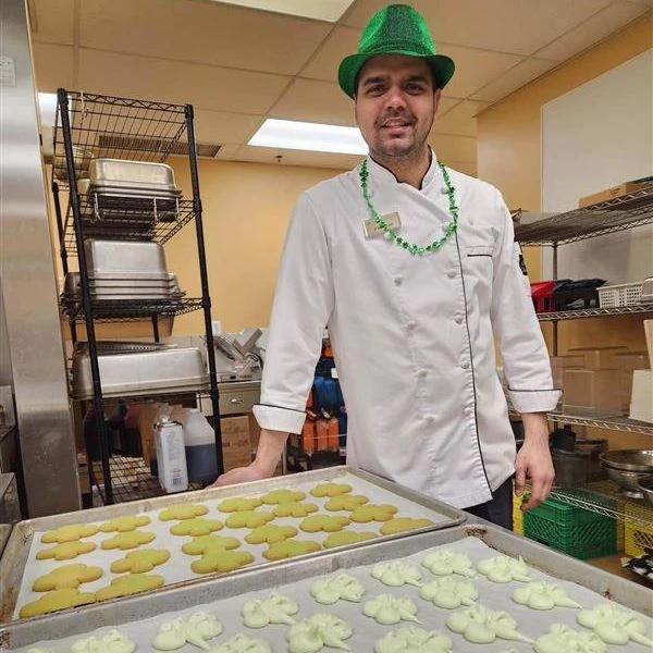 A chef standing with a green fedora on. He is next to a couple trays of cookies cut in the shape of clovers