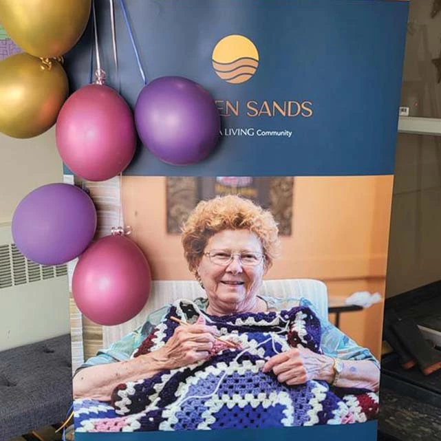 Golden Sands pull up banner featuring a senior woman knitting a blanket. Balloons of various colours are tied to the top of the banner, hanging downwards.