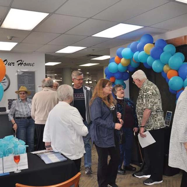 The Deer Park Village open house, with multiple people socializing. There is a large balloon arch behind them with blue, gold, teal, and orange balloons.