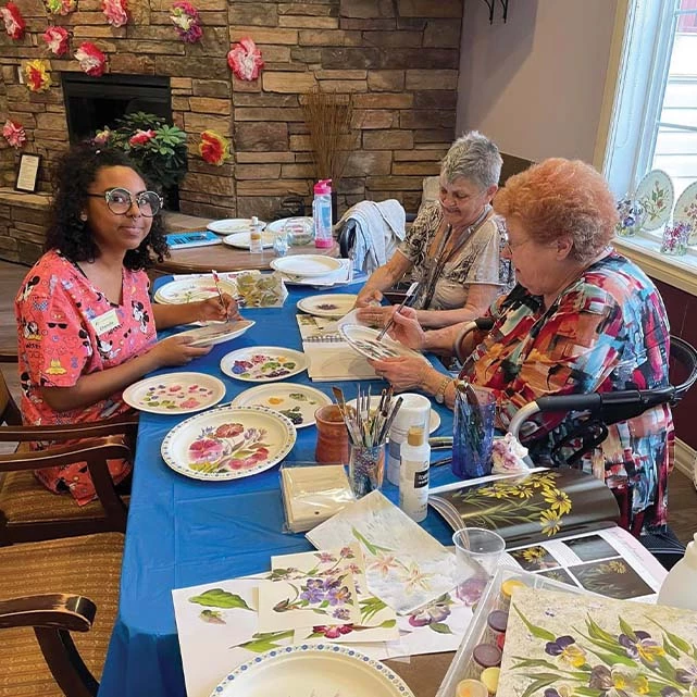 Two senior residents along with a staff member painting beautiful floral designs onto plates. Behind them is a fireplace.