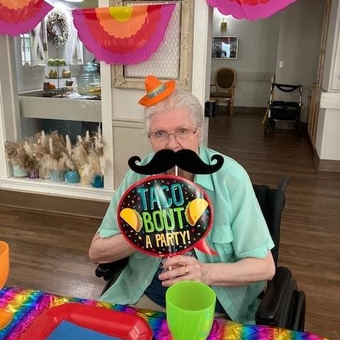 A senior celebrating Cinco de Mayo. She is holding decorations and wearing a sombrero. She also has a moustache cutout she is holding up to her face.