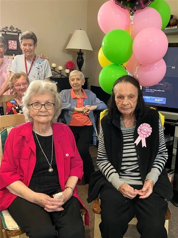 Senior residents sitting together smiling. One woman has balloons behind her and a ribbon on her shirt.