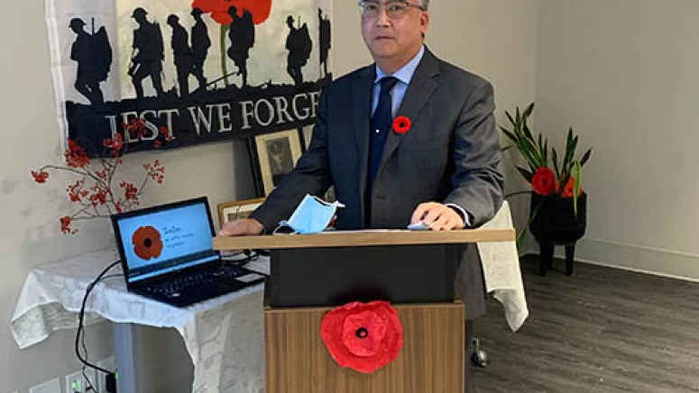 Man standing in a suit for the Remembrance Day ceremony. Behind him is a flag with a poppy and 