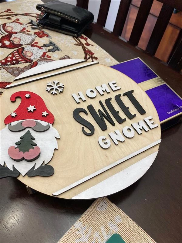 Christmas decoration on a round piece of wood. There is a gnome with a christmas hat on the sign with the words,