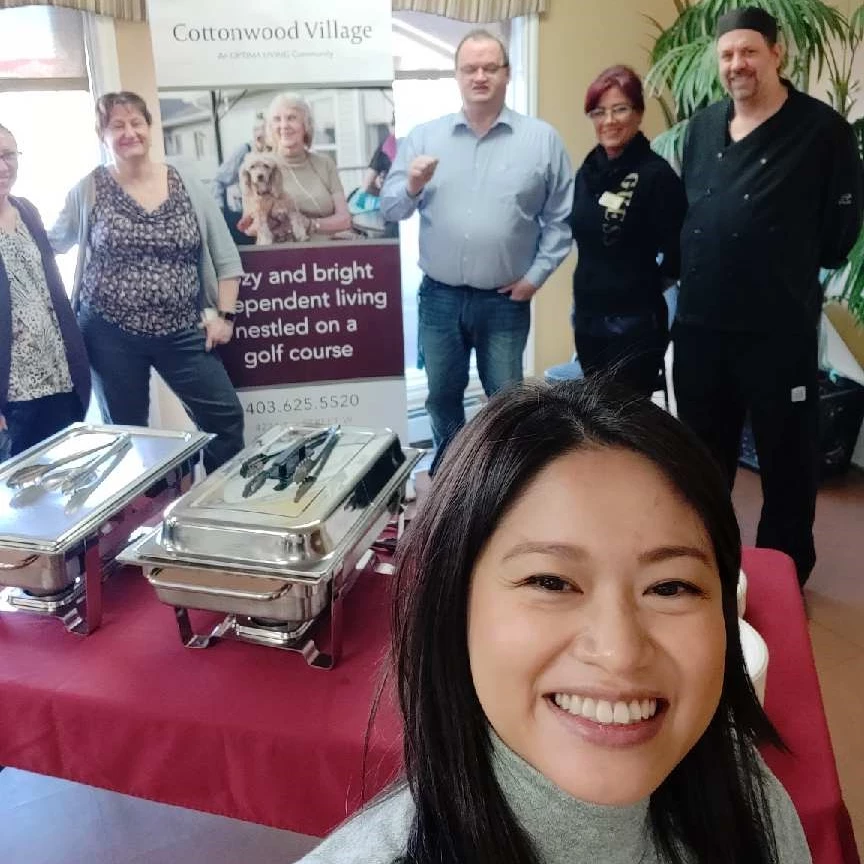 Cottonwood Village open house display. Young woman taking a selfie with other staff in the background, including the General Manager and Chef of Cottonwood Village.