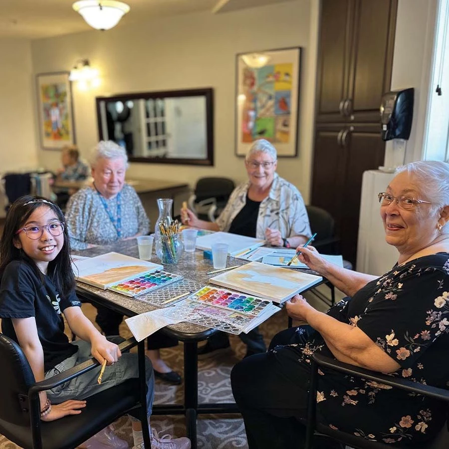 3 Senior women smiling at a table painting alongside a little girl.