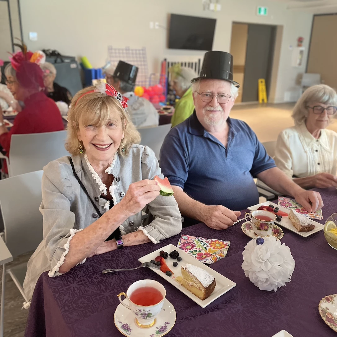 Multiple residents seated at a table with various snacks enjoying watermelon, cake, and tea. The one woman has a bow bandana on and the man has a plastic top hat.