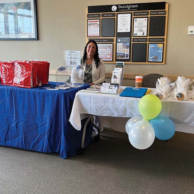 Woman seated behind some information tables at Sweetgrass. She has cookies, bags, balloons, and a prize draw in front of her.