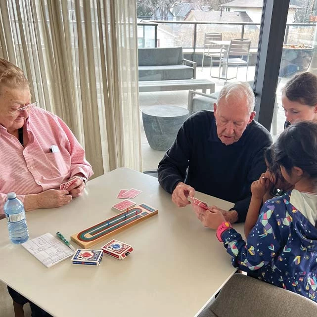 Two seniors playing cribbage with two young girls. The senior man is showing them how to play.