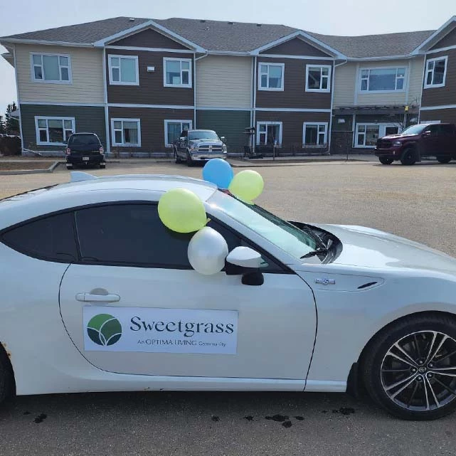 A white car with balloons and a Sweetgrass decal on the side door. The car is parked in front of the building and was decorated to celebrate the open house.