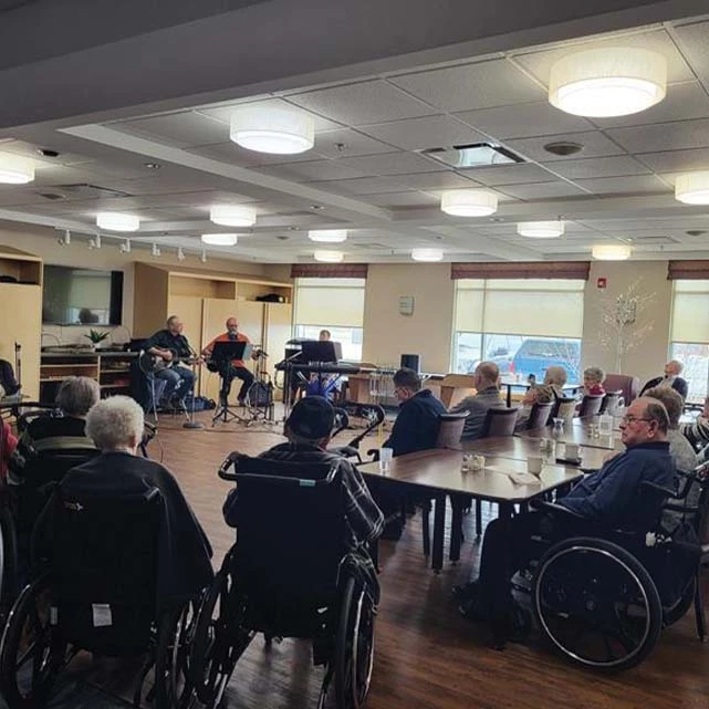 Multiple seniors seated watching and listening to a performance of three musicians. The musicians are playing a couple guitars, and a keyboard.