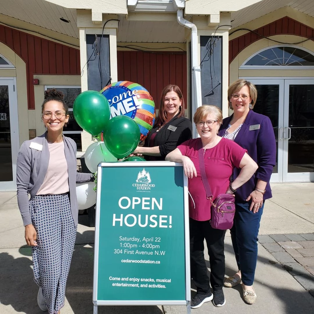 Four staff members standing together with balloons and an open house sign. The balloons are mostly green and white, along with a large balloon that says, 