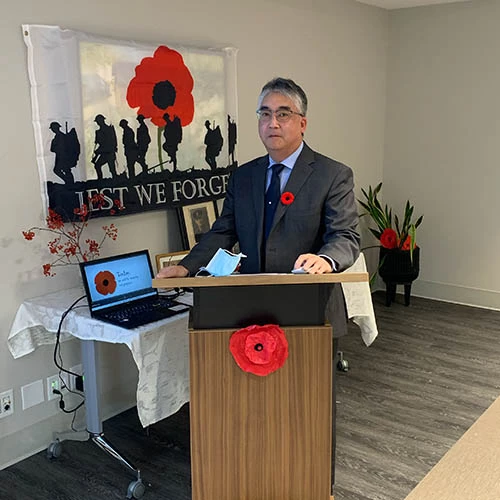 Remembrance Day service feature for Wisteria Place. Sharply dressed man in a suit is standing in front of a flag with a poppy that says, 
